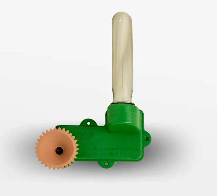 Fotografía real del alimentador con laberinto: cuerpo de plástico impreso en verde, con el tubo dosificador de vidrio montado en vertical y, a un lado, el disco dentado de barro cocido. El canal en zigzag deja entrar a las obreras sin que se ahoguen en el jarabe.
